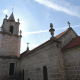 Igreja de pedra com torre sineira quadrangular, cruzes no telhado e céu azul.