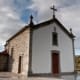 Capela de arquitectura tradicional portuguesa com fachada de pedra e cruz no topo, rodeada de paisagem rural.