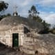 Capela construída em granito com porta verde, cruz branca no cume, rodeada por pinheiros.