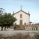 Igreja branca com cruz no topo, cemitério com sepulturas de pedra e árvore frondosa ao lado.