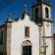 Capela de fachada branca com torre sineira, cruz no topo, porta verde e janela, em rua de pedra.