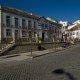 Praça pedonal com edifício azul e branco de dois pisos, telhado de cerâmica laranja, fachadas de casarões e céu limpo.