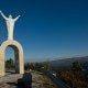 Monumento com figura humana com braços erguidos no topo de arco pétreo, vista para vale montanhoso.