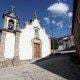 Igreja branca com dois sinos e porta vermelha, em rua de pedra, sob céu azul