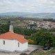Capela branca com telhado de telha vermelha em primeiro plano, vista panorâmica de aldeia com casas de telha vermelha no vale ao fundo