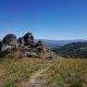 Formação rochosa granítica num monte com vegetação rasteira e vista panorâmica de vale e montanhas ao fundo.