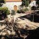 Pátio de terra com plantas e arbustos, porta arqueada amarela em parede cal, telhado cerâmico