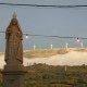 Estátua religiosa de pedra com coroa, ladeada por fios de iluminação, com aerogeradores visíveis no horizonte enevoado.