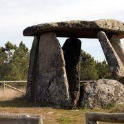 Dolmen com lápide horizontal suportada por três menhires verticais, paisagem rural ao fundo. — ampliar imagem