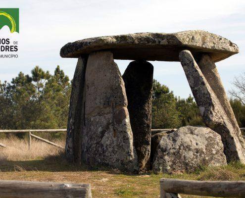 Dolmen com lápide horizontal suportada por três menhires verticais, paisagem rural ao fundo. — ampliar imagem
