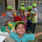 Crianças em coletes amarelo-fluorescente e bonés verdes participam em atividade educativa com materiais de construção e segurança num pátio.
