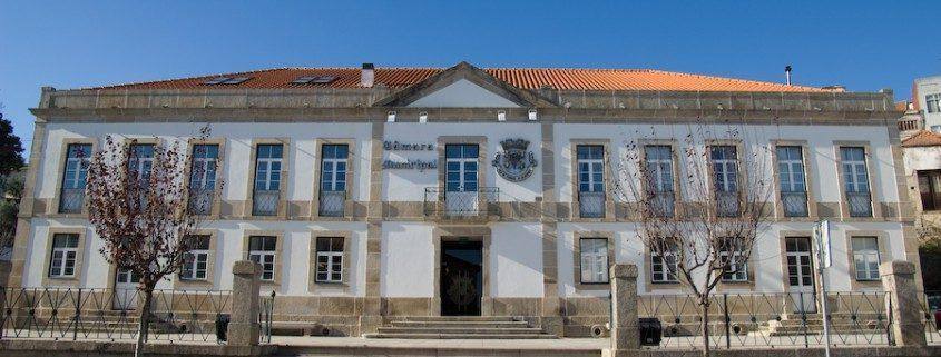 Edifício institucional de dois pisos com fachada azul e branca, escadas de acesso frontal e brasão no centro