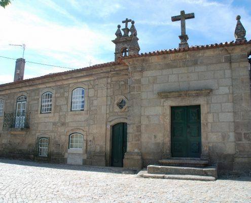 Edifício de alvenaria em pedra com fachada simples, duas portas verdes, janelas e cruz no telhado.