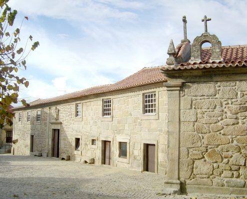Casa senhorial de pedra com telhado de cerâmica, cruzeiros no topo e pátio pavimentado.