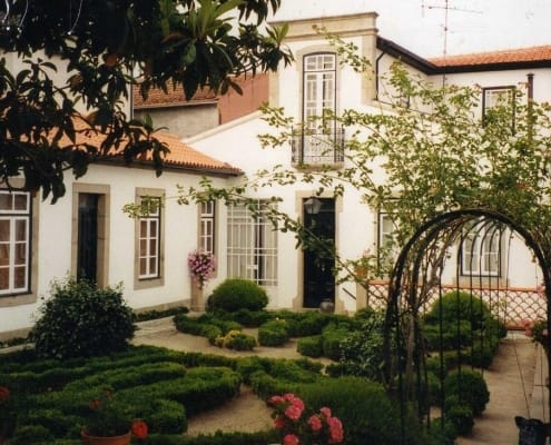 Pátio de casa colonial com arco de ferro, flores vermelhas, arbustos aparados e janelas de vidraça.