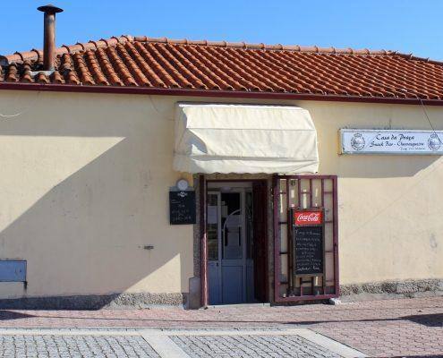 Fachada de estabelecimento com parede branca, porta vermelha, toldos brancos e telha cerâmica vermelha.