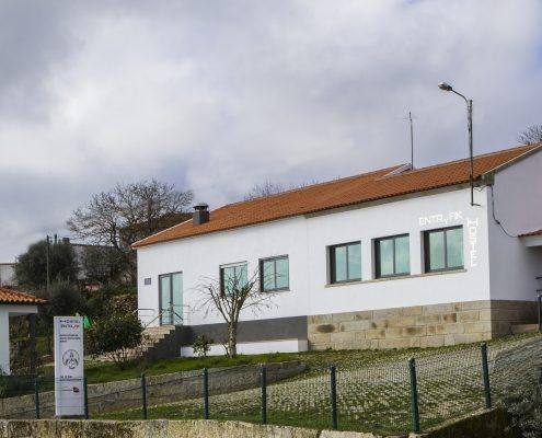 Fachada branca com telhado em telha vermelha, vedação e portão de ferro, envolvida por vegetação e céu nublado.