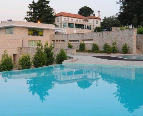 Piscina com água azul-turquesa refletindo edifício moderno de pedra e mansão histórica ao fundo