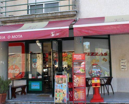 Entrada de pizzaria com toldo vermelho, janelas amplas, máquina de refrigerante e decoração temática.