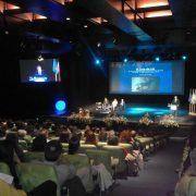 Auditório cheio com público sentado a assistir a painel de discussão no palco, com ecrãs de projeção e iluminação azul.