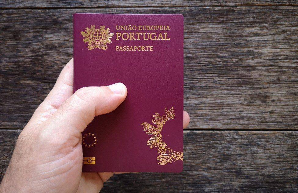 Passaporte português de capa vermelha-escura com brasão e inscrições em ouro, seguro numa mão contra fundo de madeira