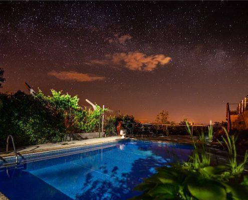 Piscina iluminada à noite com vegetação circundante e céu estrelado ao fundo.