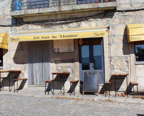Fachada de café com toldo amarelo, cadeiras pretas e porta azul numa rua em calçada portuguesa