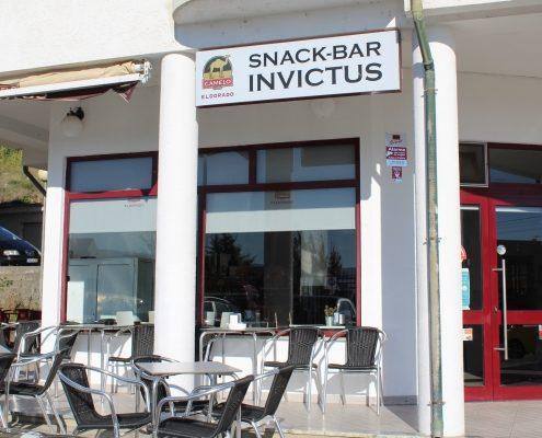 Fachada branca e vermelha do Snack-Bar Invictus com esplanada de mesas e cadeiras cinzentas.