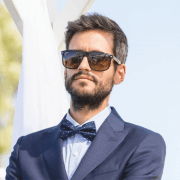 Homem com barba, óculos de sol castanhos, fato azul-marinho e laço azul com pontinhos brancos