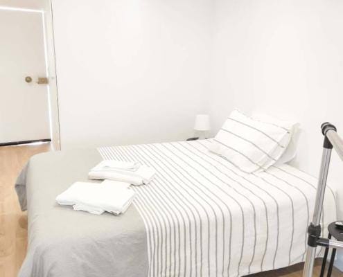 Quarto minimalista com cama de casal, roupa de cama branca com padrão de riscas, almofadas e mantas dobradas.