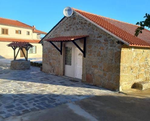 Pequena casa de pedra com telhado de telha cerâmica, antena parabólica no cume e pórtico de madeira, num pátio empedrado.
