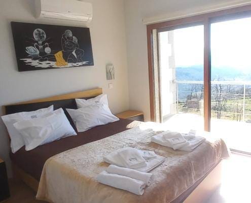 Quarto com cama de casal, mesinha lateral, quadro decorativo preto na parede, ar condicionado e porta de vidro com vista para paisagem montanhosa.