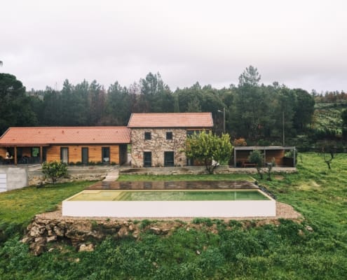 Casa moderna com fachada em pedra e laranja, piscina branca, envolvida por vinhas e floresta