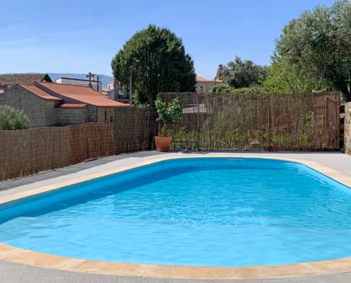 Piscina azul redonda com deck em cimento, cercada por vedação em palha e árvores, edifícios rurais ao fundo.