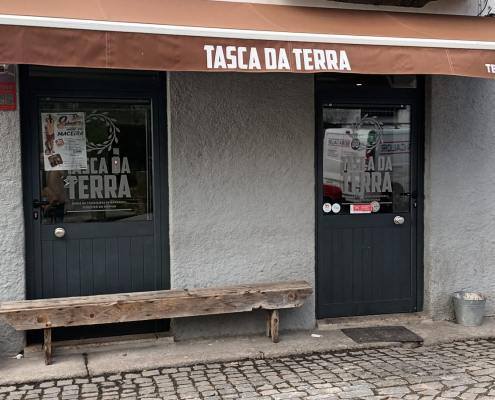 Fachada de estabelecimento com toldo castanho, nome Tasca da Terra em branco, porta preta dupla e janelas pequenas, parede branca, piso em calçada portuguesa.