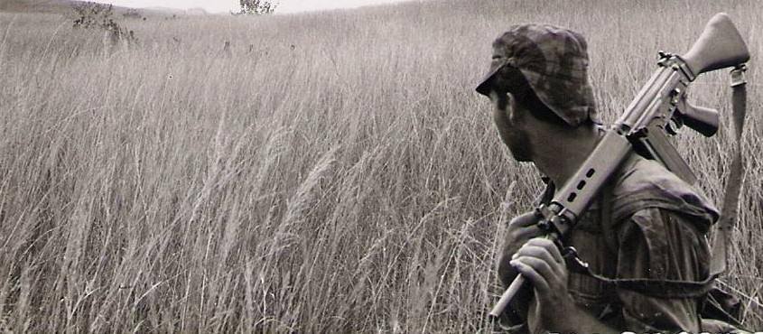 Soldado em pé num campo de vegetação seca, segurando arma de ombro com luneta, de perfil.