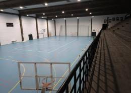 Interior de pavilhão desportivo com piso azul, marcações de linhas brancas e vermelhas, redes de futsal, iluminação LED e bancadas laterais.