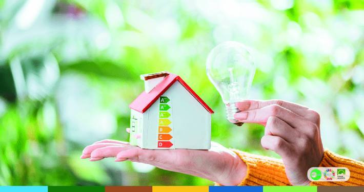Mão segurando casa miniatura com rótulo de eficiência energética e lâmpada acesa contra fundo verde desfocado