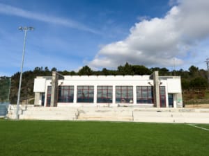 Edifício moderno de piscinas com fachada branca, grandes janelas e piscina exterior de relva sintética.