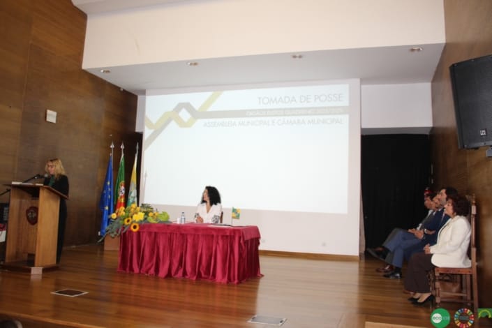 Cerimónia de tomada de posse com oradora ao podium, painel presidencial e assistentes sentados numa sala com bandeiras.