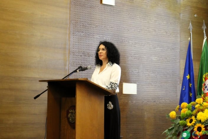 Mulher de óculos ao pódio com microfone numa sala de conferências com bandeiras oficiais.