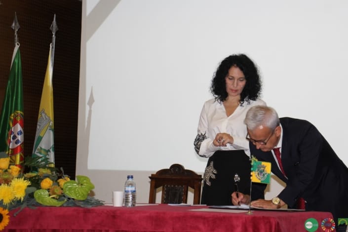 Homem de fato escuro e mulher de blusa branca em cerimónia oficial com bandeiras portuguesas ao fundo.