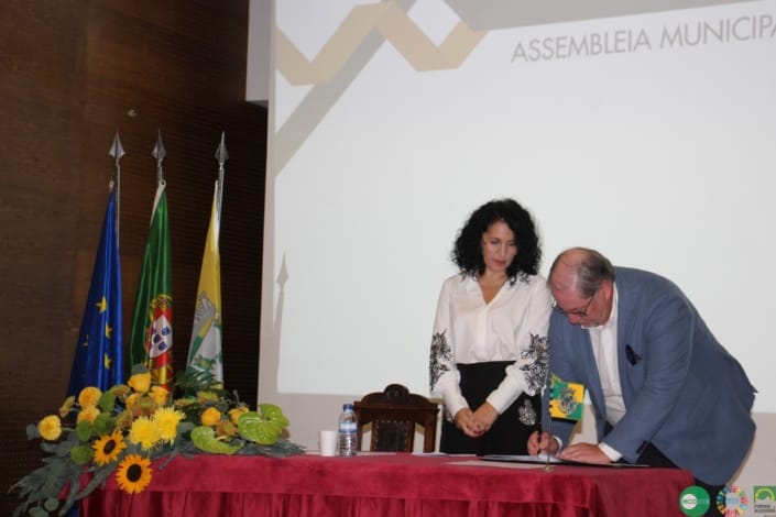 Dois representantes em cerimónia oficial assinam documento junto a bandeiras e flores amarelas.