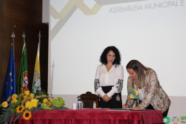 Duas mulheres numa sessão oficial da Assembleia Municipal, junto a mesa com flores amarelas e bandeiras institucionais.