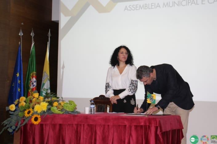 Homem de fato preto assina documento em mesa enquanto mulher o observa.