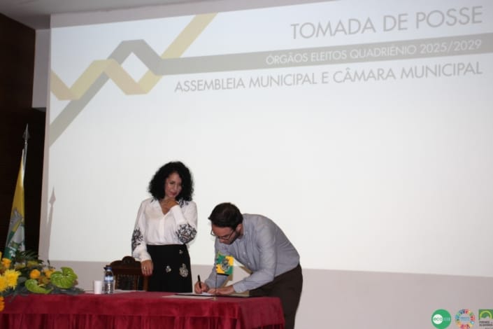 Dois autarcas na assinatura de documentos durante cerimónia de tomada de posse, com projeção institucional ao fundo.