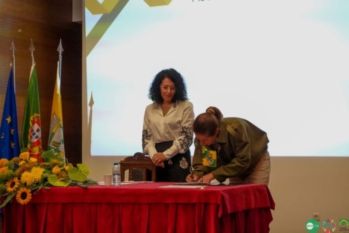 Duas mulheres em cerimónia oficial: uma de pé junto a bandeiras, outra assinando documento sobre mesa com toalha vermelha.