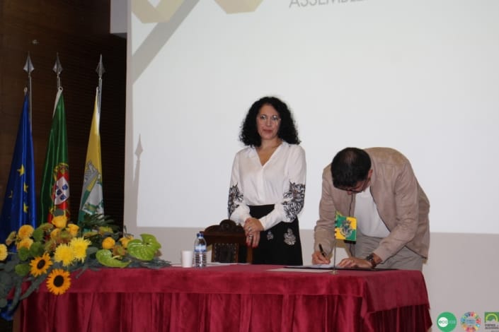 Duas mulheres em sessão solene perante três bandeiras e flores amarelas numa mesa com toalha vermelha.