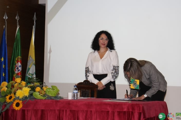 Duas mulheres numa mesa oficial com bandeiras portuguesas, durante uma sessão de trabalho com assinatura de documento.
