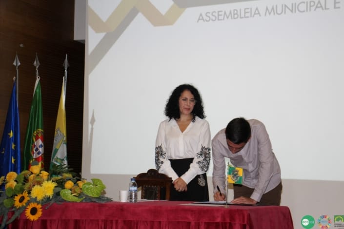 Duas pessoas em cerimónia oficial junto a mesa com bandeiras e flores